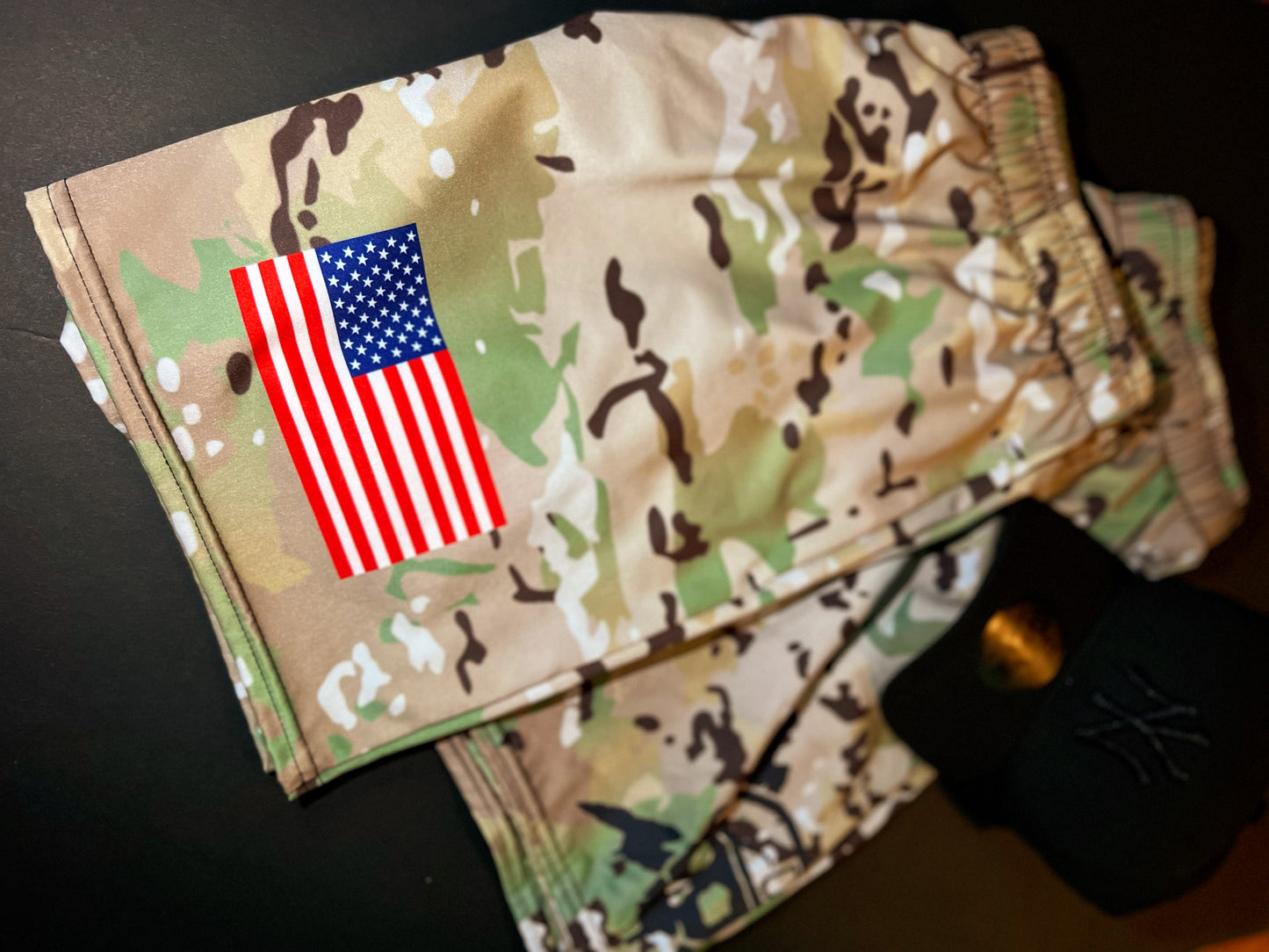 Camouflage shorts in a tan, green, and brown pattern with a printed American flag graphic on the left leg, shown on a dark surface next to a black baseball cap.