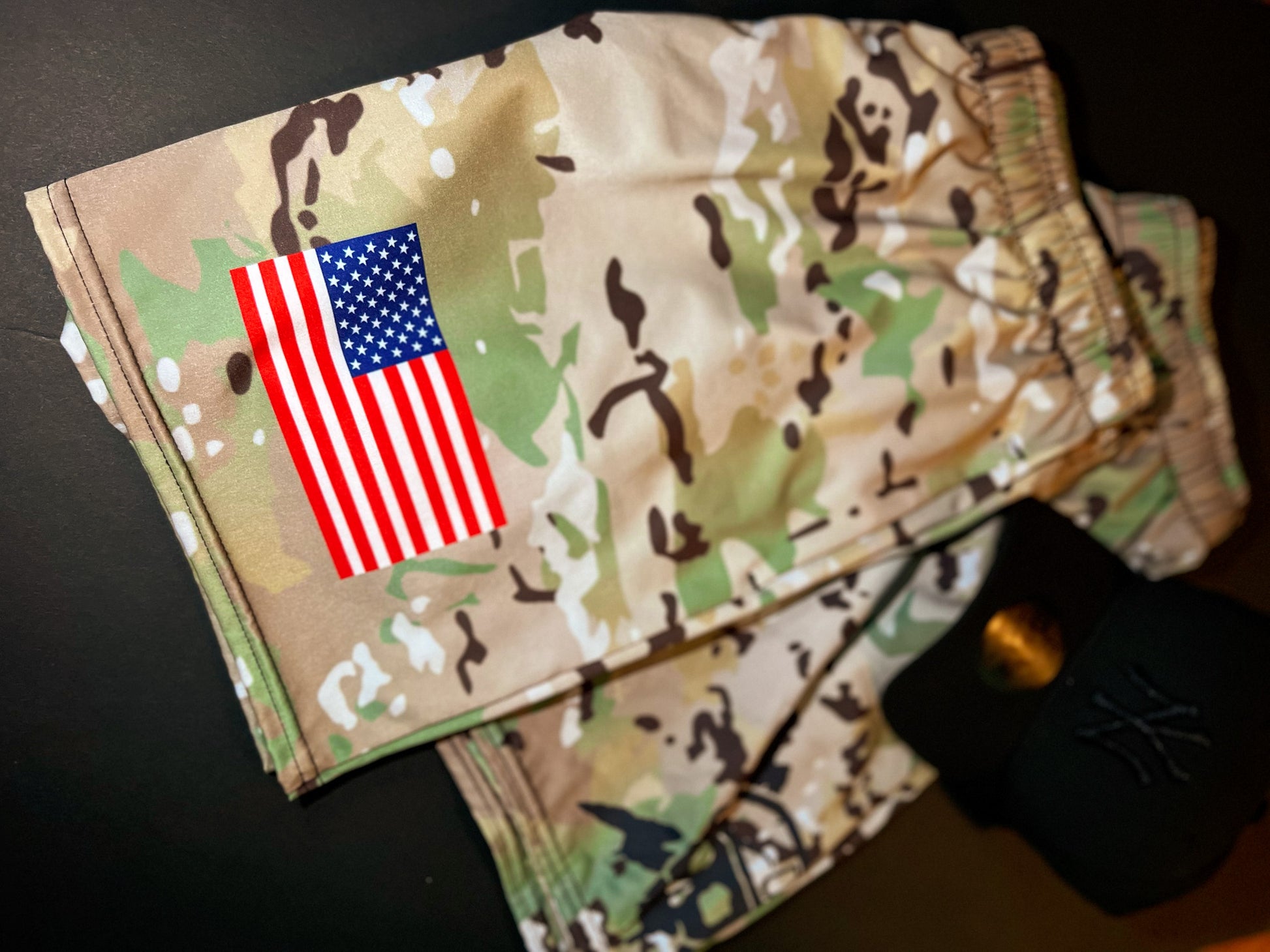 Camouflage shorts in a tan, green, and brown pattern with a printed American flag graphic on the left leg, shown on a dark surface next to a black baseball cap.